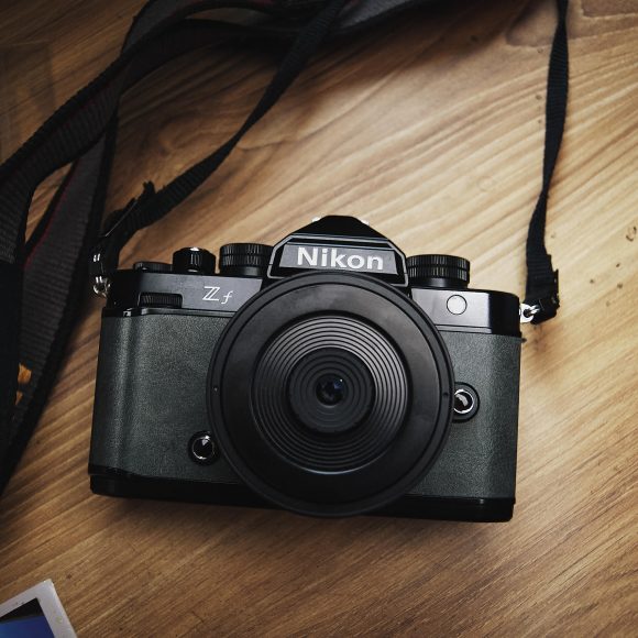 A Nikon Z fc camera with a wide lens sits on a wooden surface, strap and photo prints nearby.