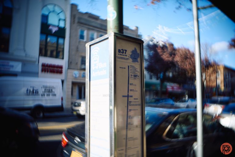 A blurry photo of a B37 bus stop sign on a busy city street with cars and buildings in the background.