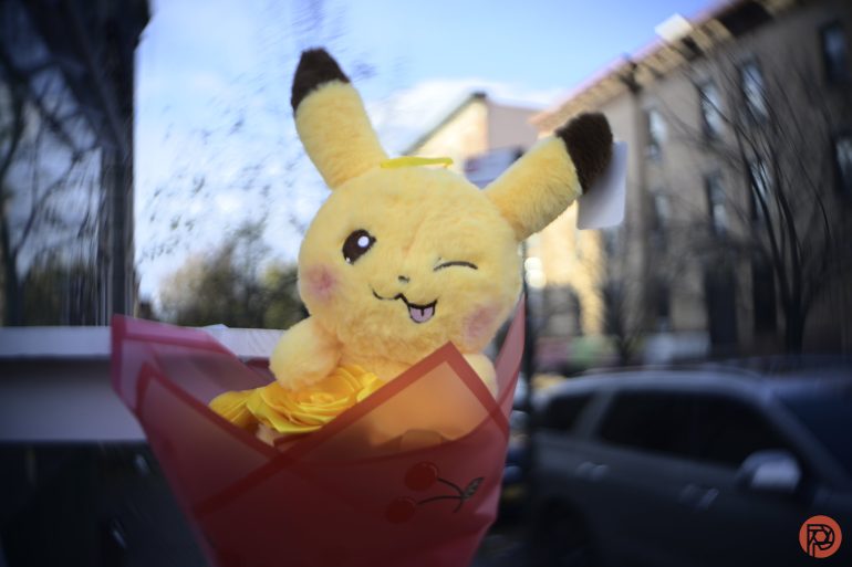 A plush Pikachu winking, wrapped in red paper, is held outdoors with buildings and cars in the background.