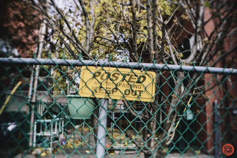 Yellow Posted No Trespassing Keep Out sign on a green chain-link fence with trees and buildings behind.