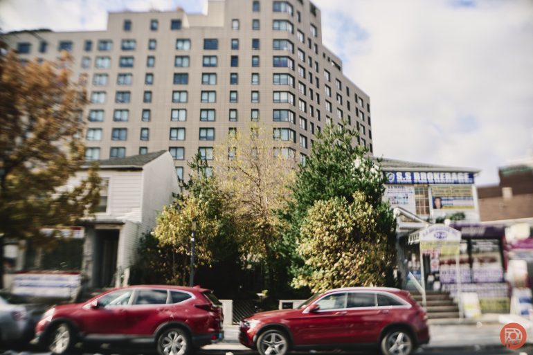 Two red cars parked on a city street with trees and a tall apartment building in the background.