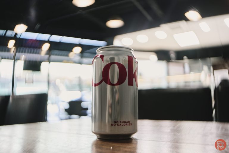 A can of Diet Coke on a wooden table in a modern, well-lit café with blurred background.
