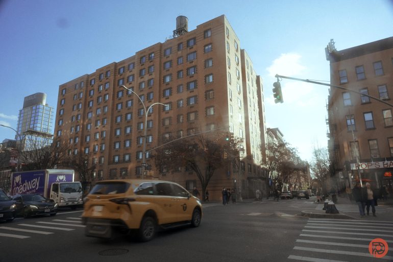 A yellow taxi drives through a city intersection beside a tall apartment building on a sunny day.