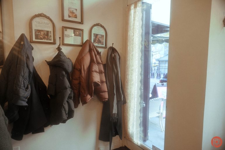 Four coats hang on wall hooks next to a window with lace curtains in a cozy, well-lit corner.