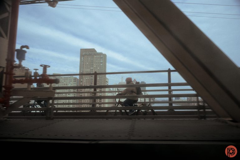 A person rides a bicycle across a bridge with city buildings and a blue sky in the background.