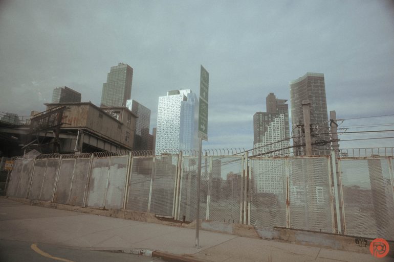 Barbed-wire fence and old building in front of modern city skyscrapers under a cloudy sky.
