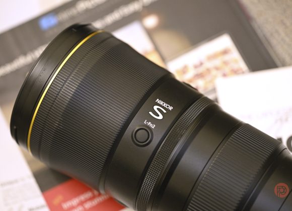 Close-up of a NIKKOR S camera lens resting on blurred papers and documents in the background.