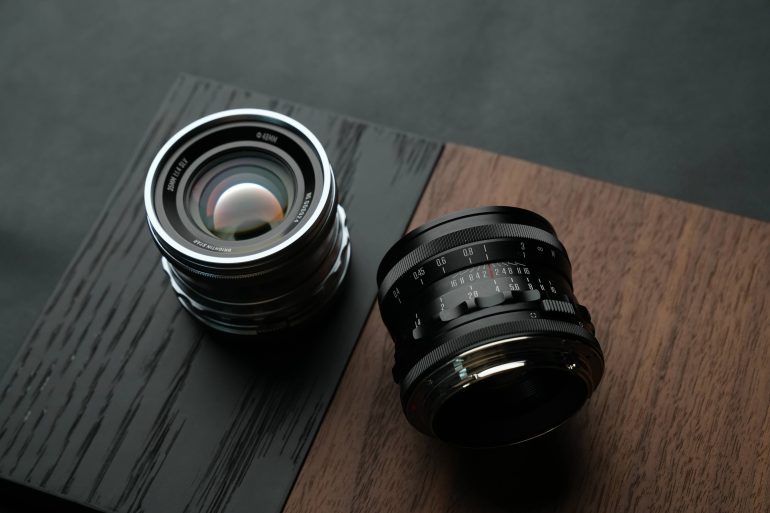 Two camera lenses rest on a two-tone wooden surface, one upright and one lying on its side.