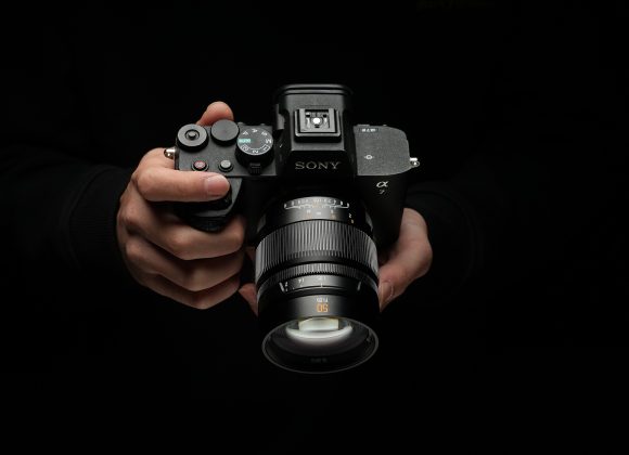 A person holding a Sony Alpha camera with a 50mm lens against a dark background.