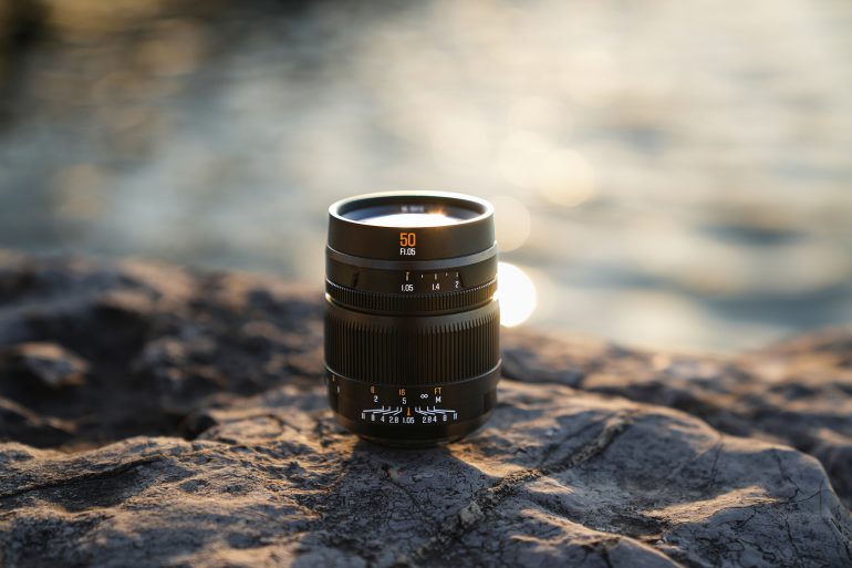 A camera lens sits on a rocky surface with blurred water and sunlight in the background.