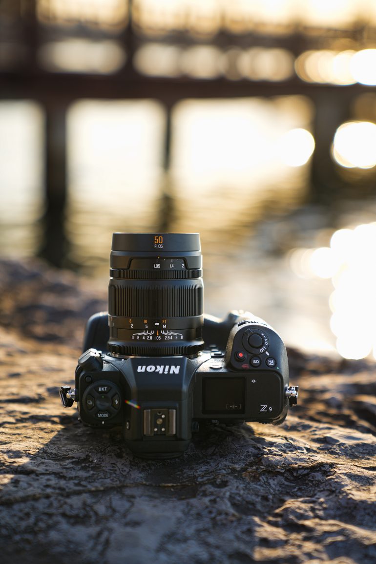 A Nikon Z camera with a 50mm lens rests on a rocky surface by the water at sunset.