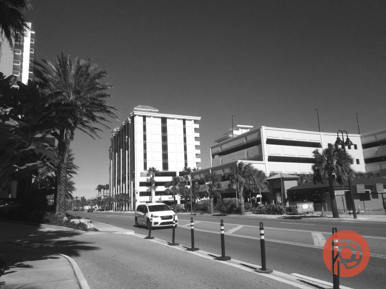 A white car drives past tall palm trees and modern buildings on a sunny city street.