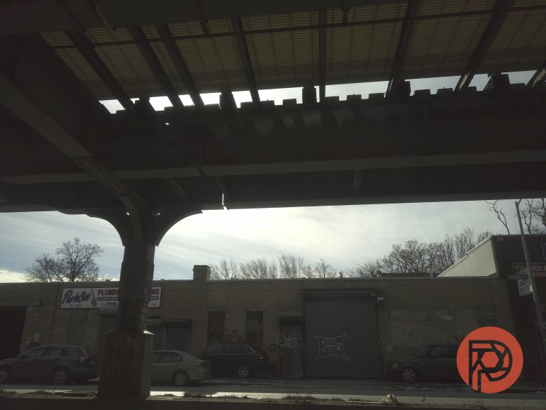 Shadowed underpass with a row of parked cars and a brick building, trees, and sky in the background.