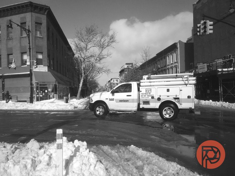A Verizon utility truck drives through a snowy city street lined with buildings under a partly cloudy sky.