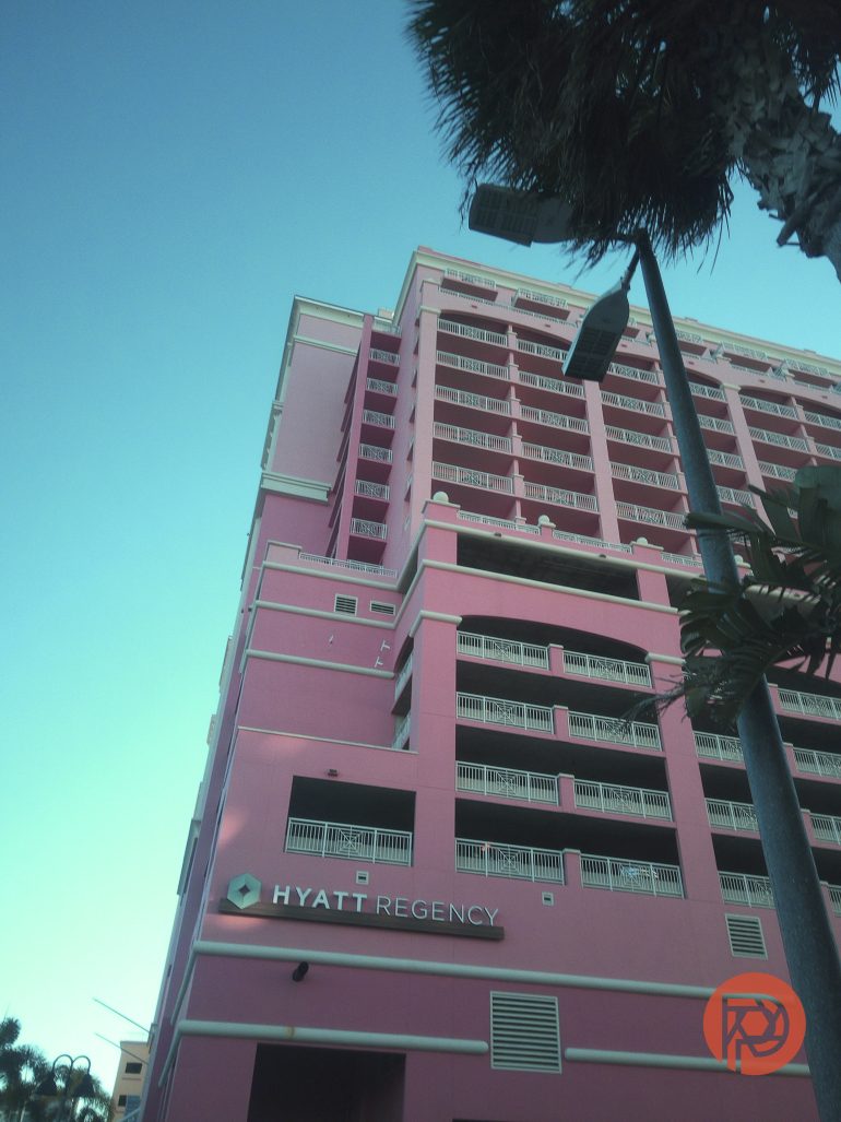 Tall pink Hyatt Regency hotel building with balconies, palm tree, and streetlight against a clear sky.