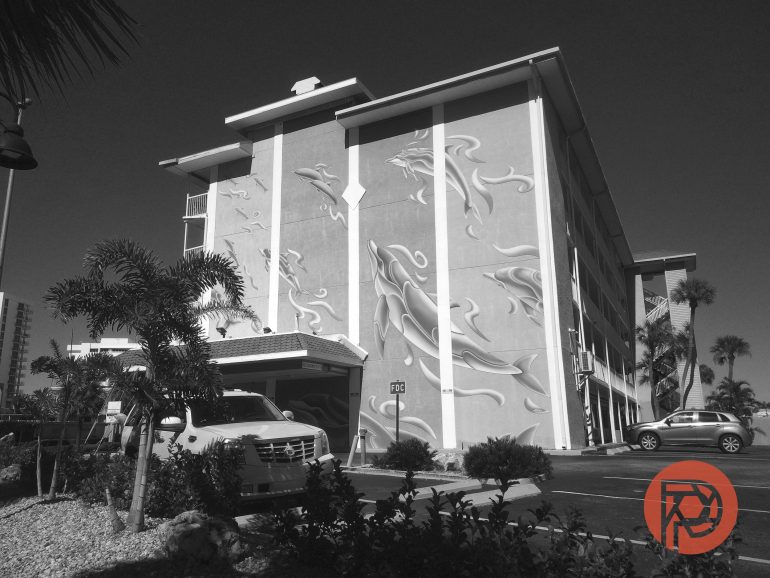 A four-story hotel with wave murals on the wall, palm trees, and cars parked in front under a clear sky.