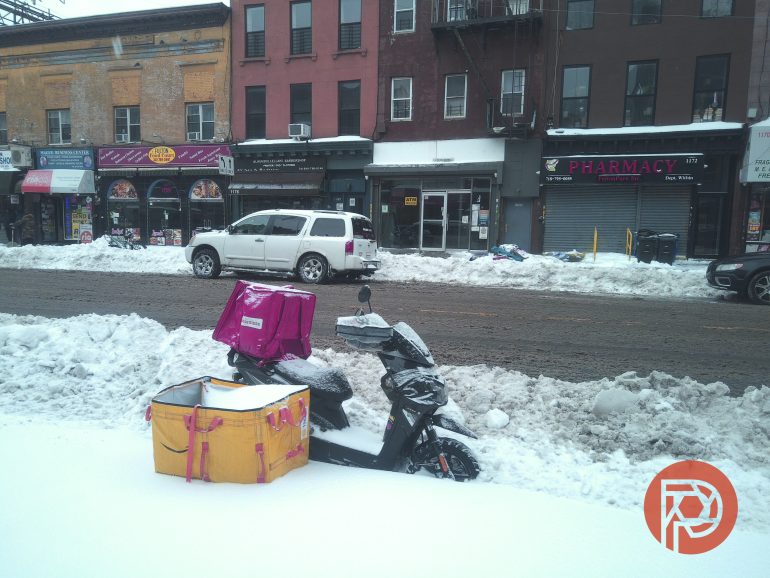 A scooter with pink and yellow delivery boxes is parked on a snowy street in front of shops.