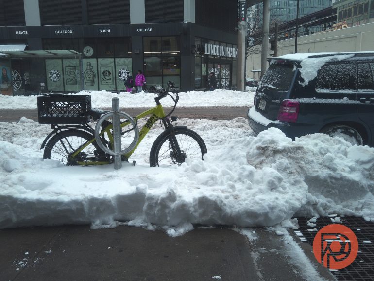 A bicycle locked to a rack is surrounded by snow on a city street with parked cars and shops in the background.