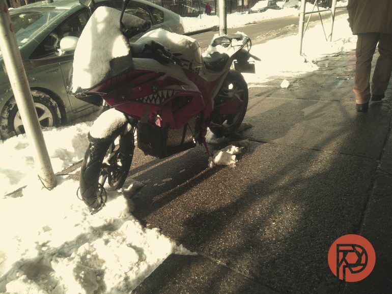 A red motorcycle with a shark mouth design is covered in snow and parked on a snowy sidewalk.