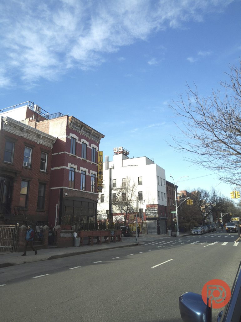 City street with brick and white buildings, leafless trees, and a clear blue sky on a sunny day.