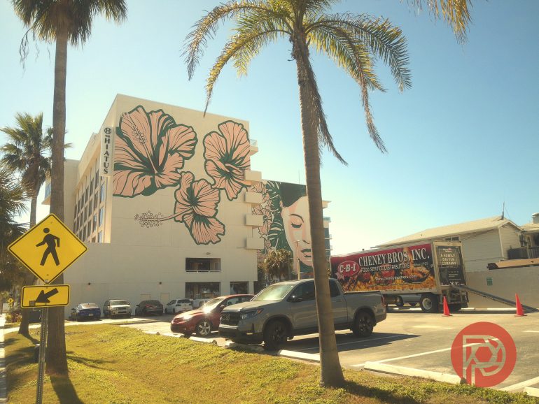 A hotel with a large floral mural, palm trees, and parked cars under a sunny sky.