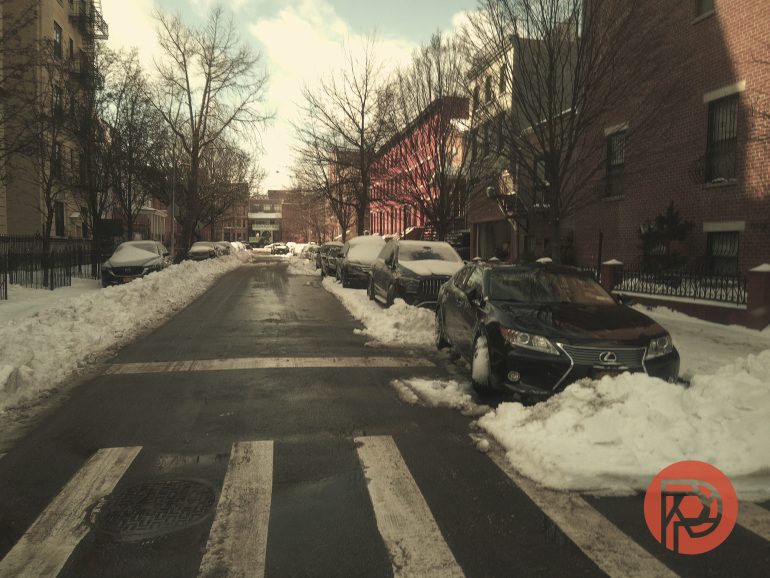 Snow-covered cars line both sides of a residential street with cleared sidewalks and bare trees.
