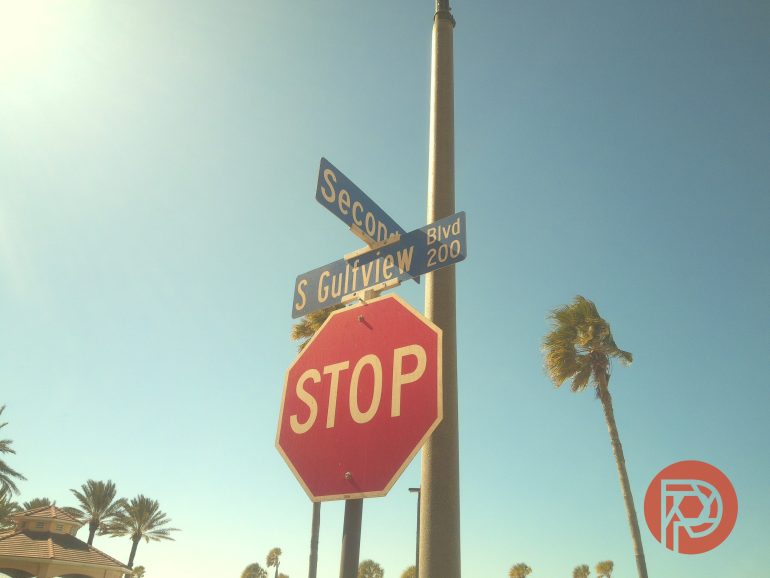 A stop sign and street signs for S Gulfview Blvd and Second St, with palm trees and a clear blue sky.