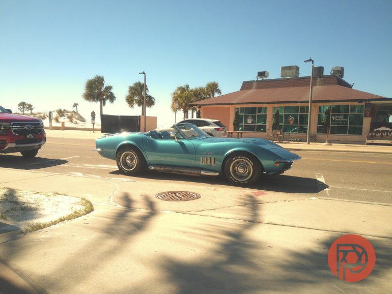 A turquoise vintage convertible drives down a sunny beachside road with palm trees and a café in the background.