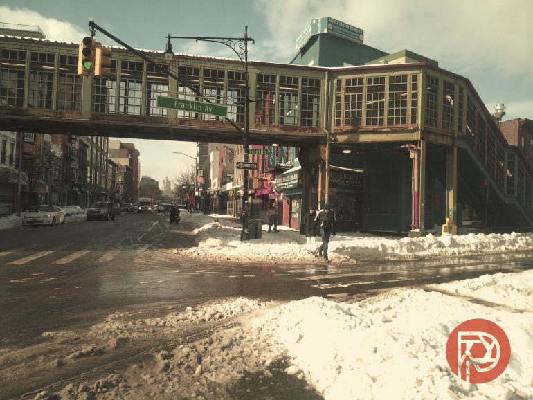 Snowy street scene with an elevated subway station at Franklin Ave, people walking, and cars in Brooklyn, NYC.