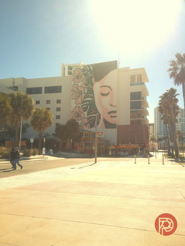 A large mural of a serene woman with flowers on a building, palm trees and pedestrians in the foreground.