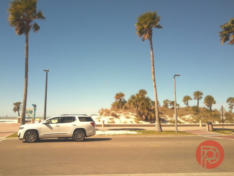 A white SUV parked by a beach road with palm trees and clear blue sky in the background.