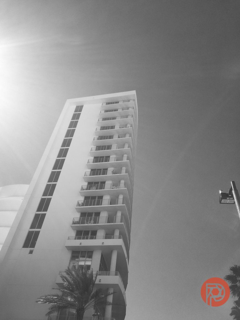 A tall, modern building with balconies in sunlight, viewed from below against a clear sky.
