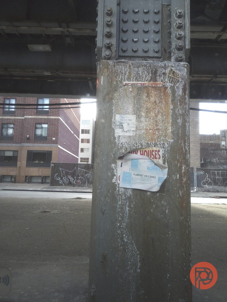 A rusty beam under a bridge with a torn paper ad, city buildings and graffiti in the background.