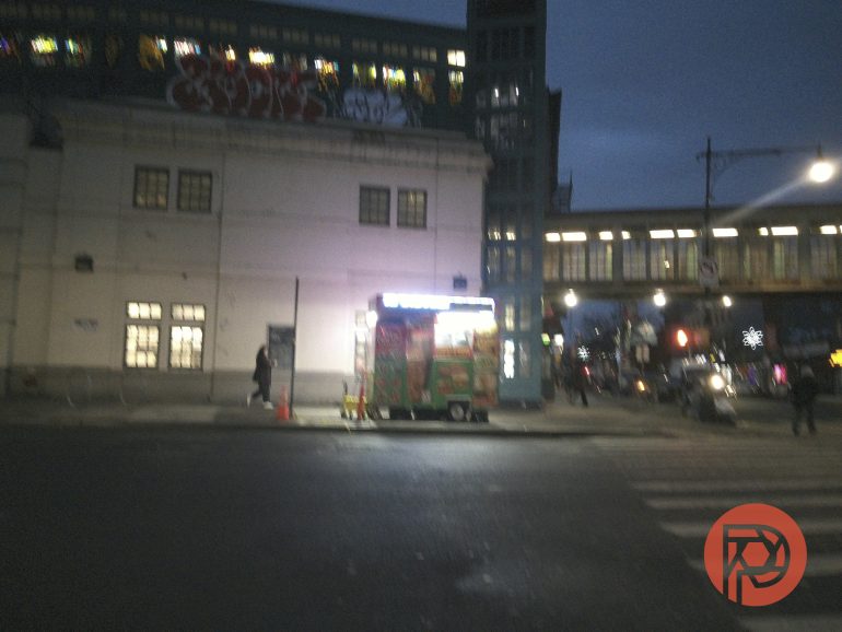A food cart with bright lights is parked on a city street at dusk near a large building and elevated train tracks.