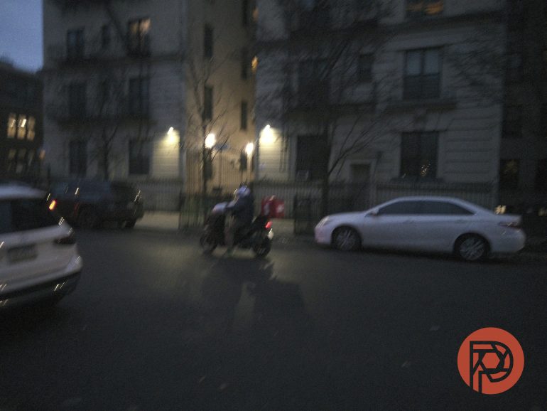 A person on a motorbike rides past parked cars on a dimly lit city street at dusk.