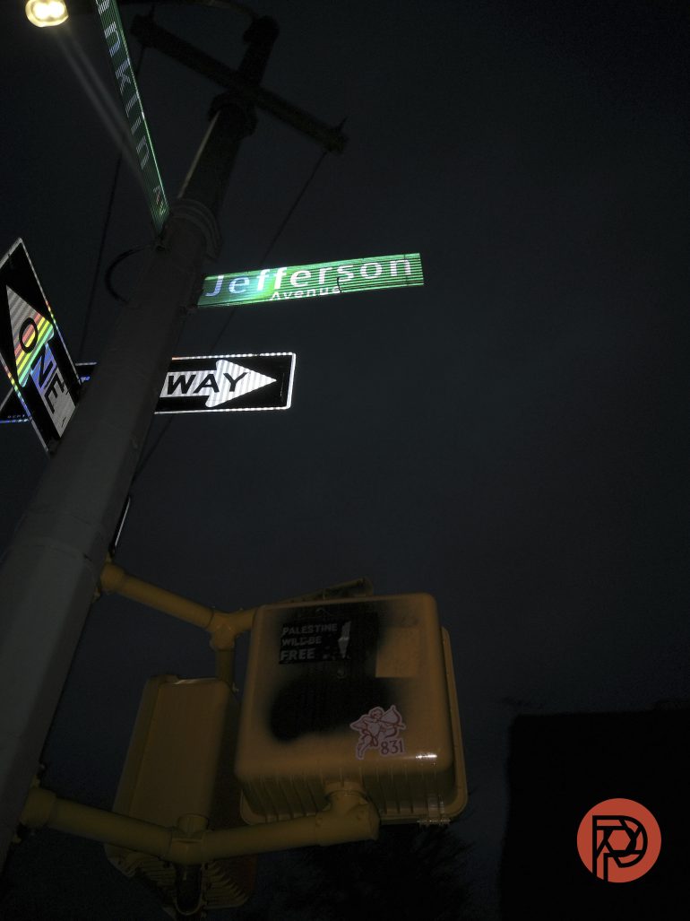 Street signs for Jefferson Avenue and One Way at night, with a sticker-covered traffic signal box below.