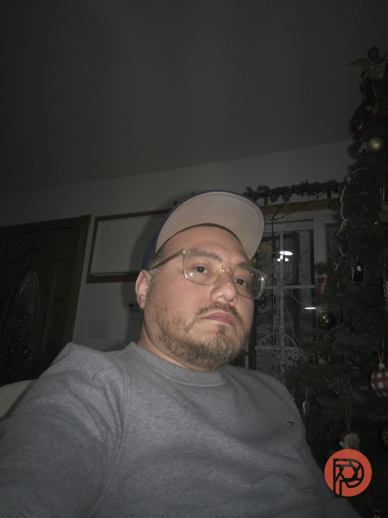 A man in glasses and a cap sits indoors near a decorated Christmas tree, looking at the camera.