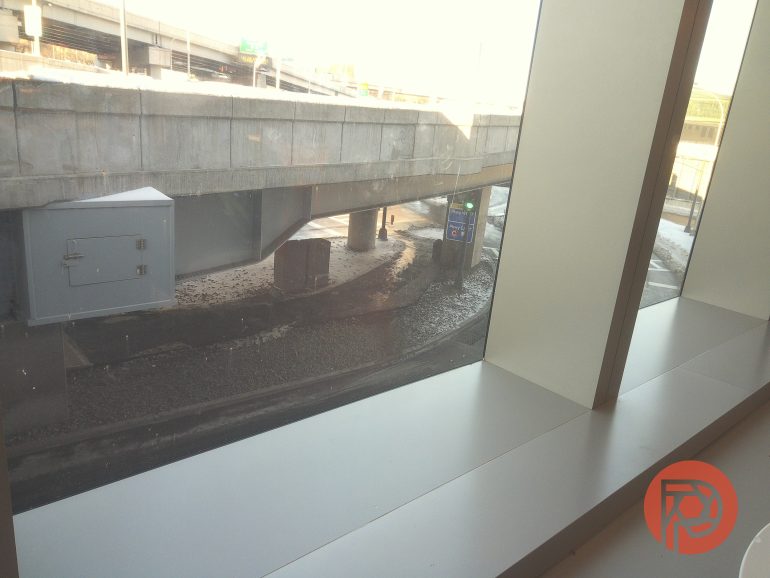 View through a window of an empty street under a highway overpass, with sunlight streaming in.