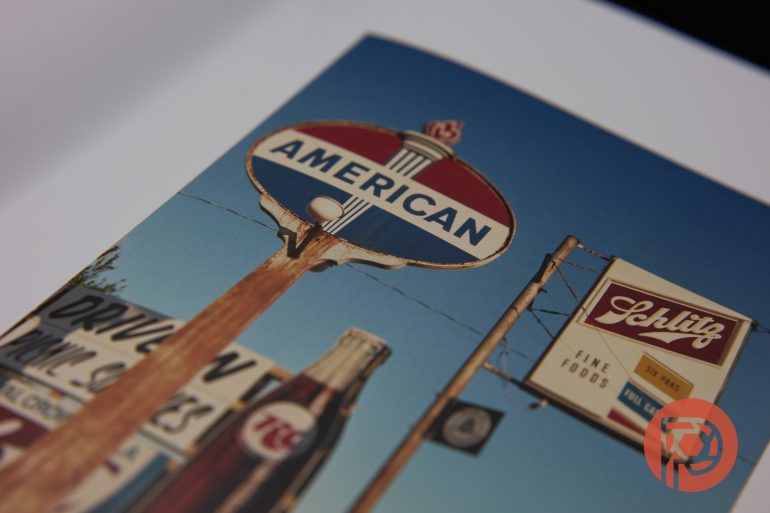 Vintage American gas station and Schlitz beer signs against a blue sky, with a blurred bottle in the foreground.