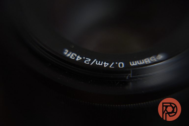 Close-up of a camera lens with white text and a red circular logo in the bottom right corner.