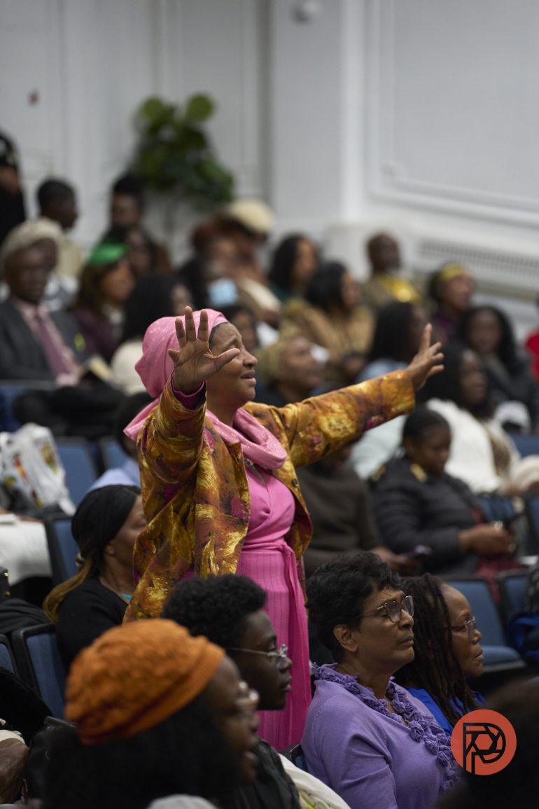 A woman in a pink dress stands with arms raised in a crowded auditorium during an event.