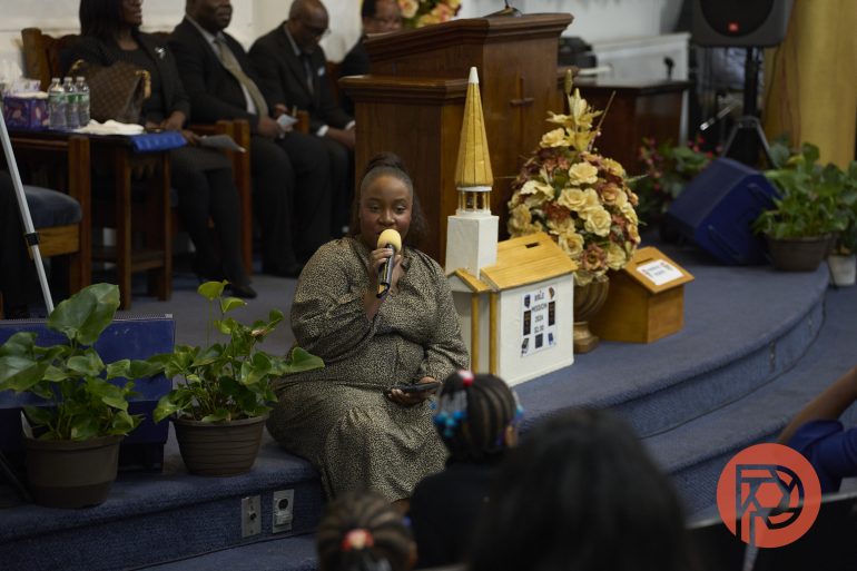 A woman sits on a church stage, speaking into a microphone to children seated in front of her.