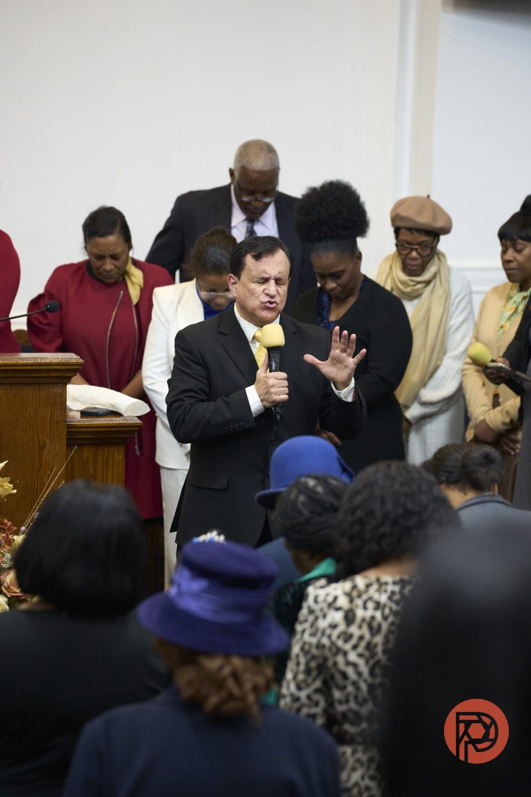 A man in a suit prays with a group of people standing behind him in a church setting.