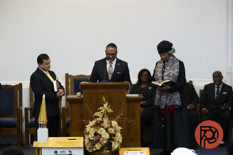 Three people stand at a church pulpit; one speaks, another reads, and a few others sit and observe.