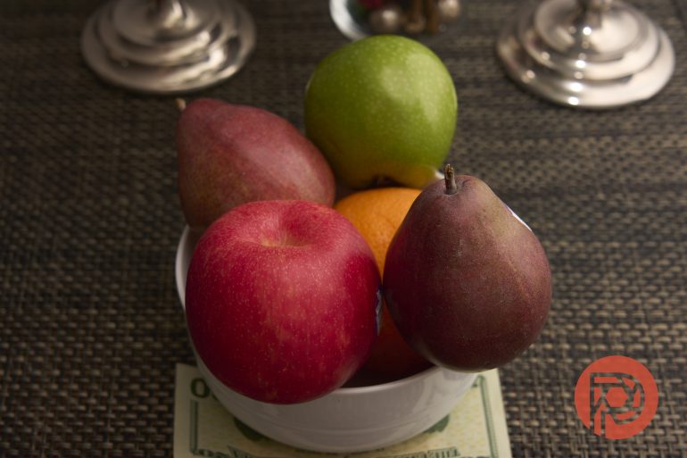 A white bowl with apples, pears, and an orange sits on a placemat with money underneath.
