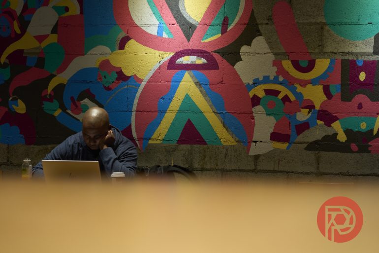 A person works on a laptop in front of a colorful mural on a brick wall.