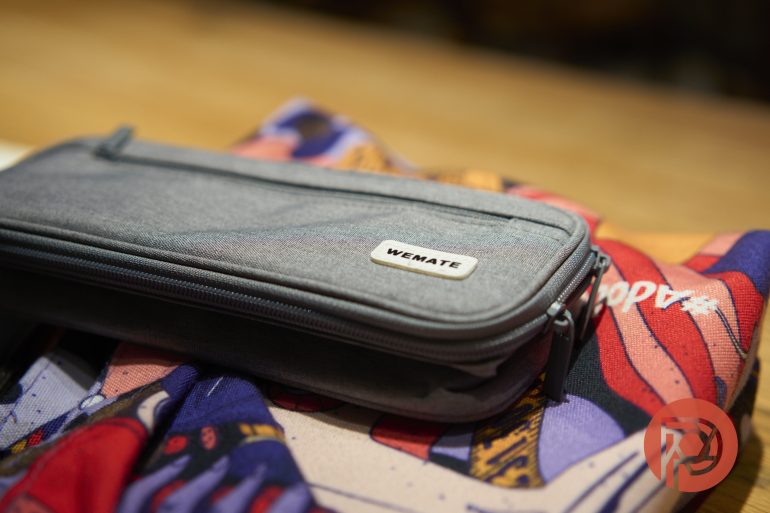 A gray WEMATE pouch rests on a colorful, patterned fabric with red and purple designs.