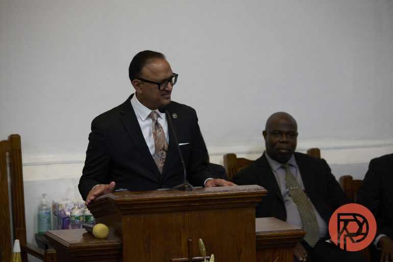 A man in a suit speaks at a wooden podium; others are seated behind him in a church setting.