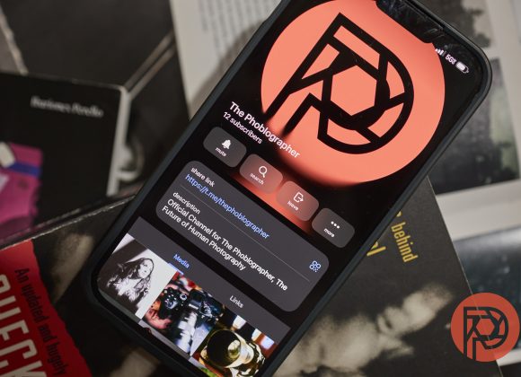 A smartphone displays The Phoblographer’s profile; books and a red logo are visible around it.