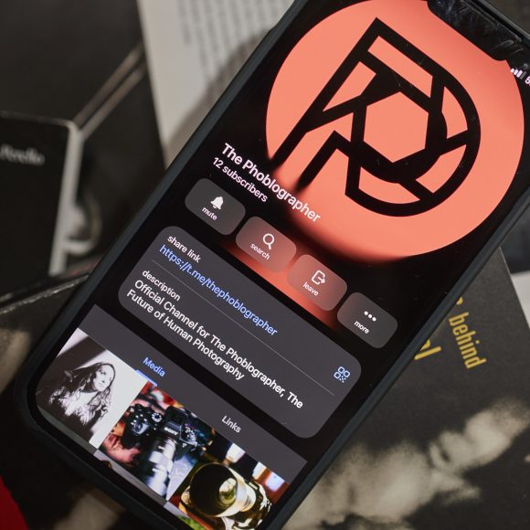 A smartphone displays The Phoblographer’s profile; books and a red logo are visible around it.